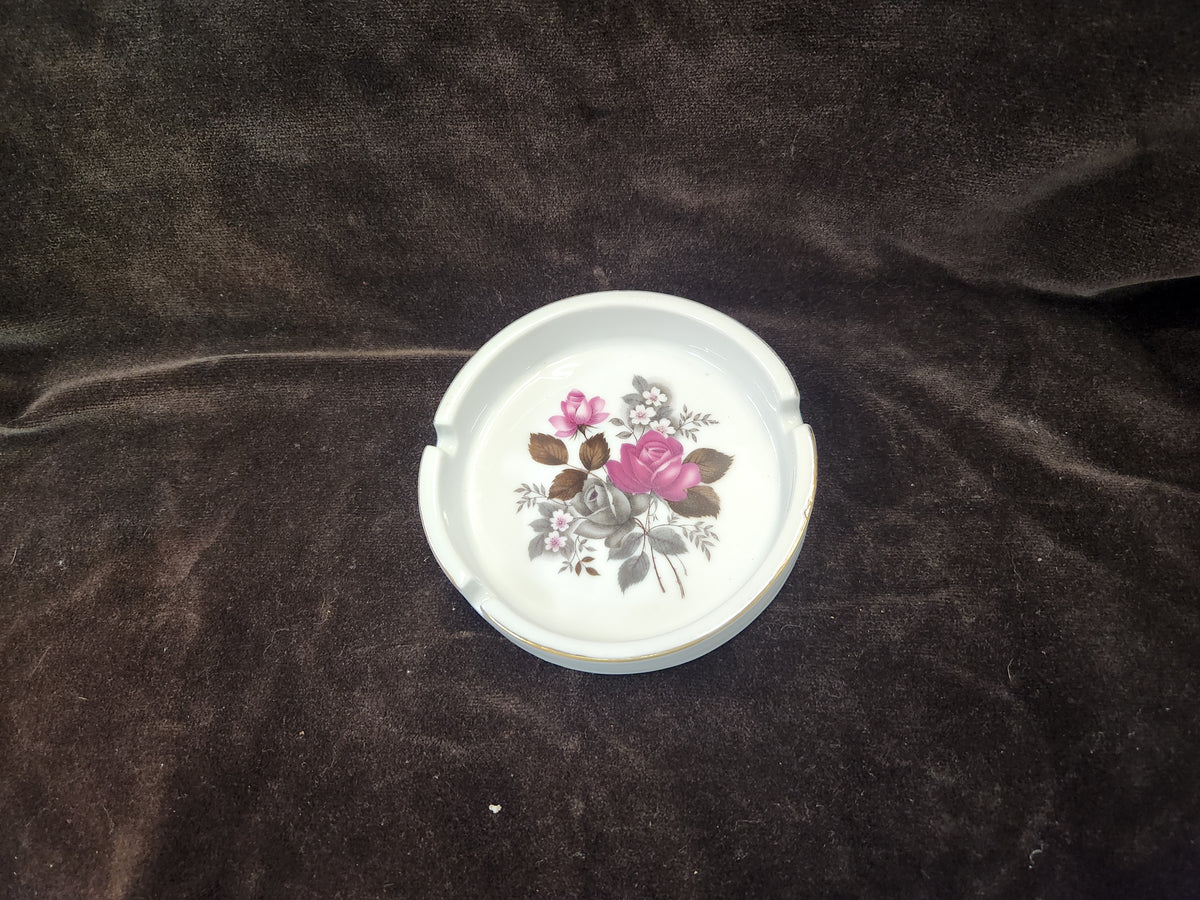 A white ceramic ashtray with a vintage floral design featuring pink roses and green leaves, set against a dark brown textured fabric background.